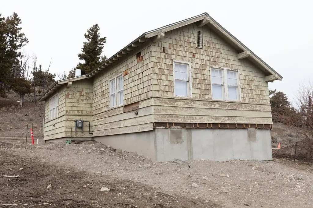 A small shingled house standing on a concrete foundation, with bare ground around it