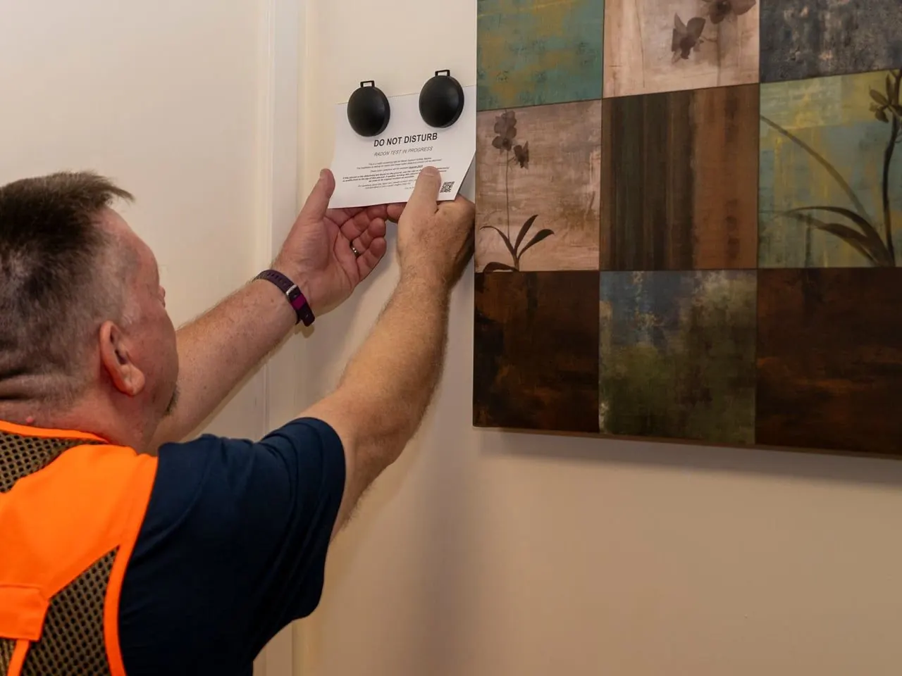 A technician in a high-visibility vest holds a card reading "Do not disturb, radon test in progress" under a pair of radon detectors on a wall