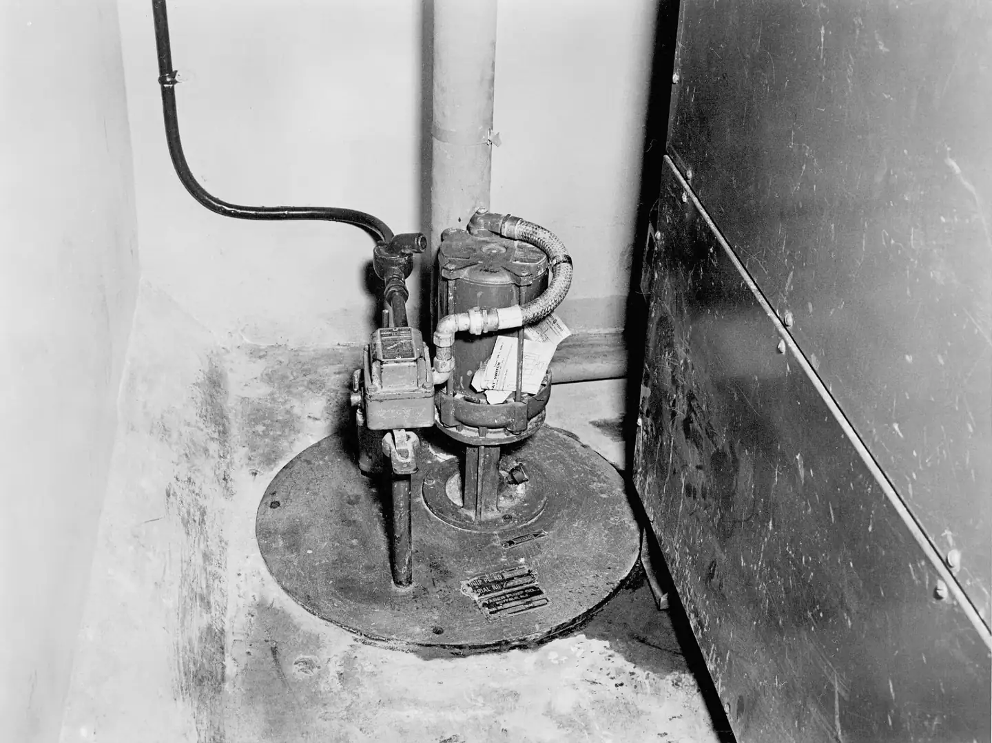 A sump pump standing on a covered pit in the corner of a concrete floor, in black and white